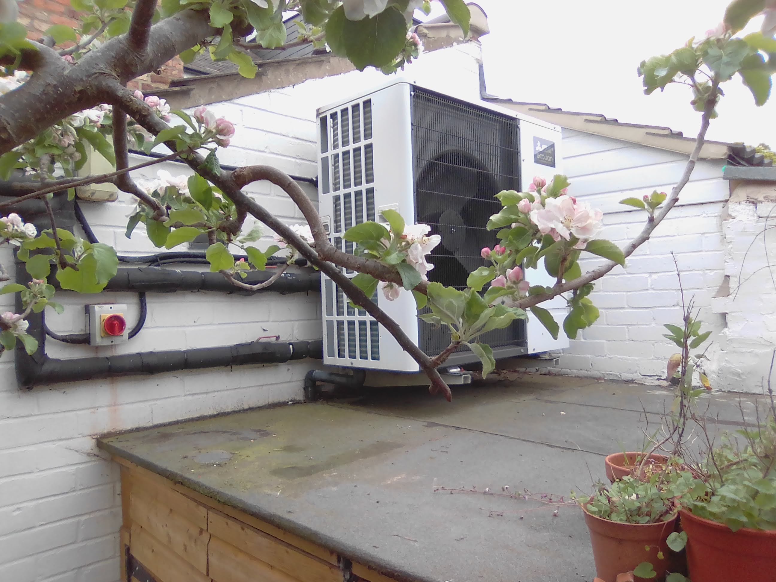 Photo of heat pump on external wall of a house with blossom in the foreground.