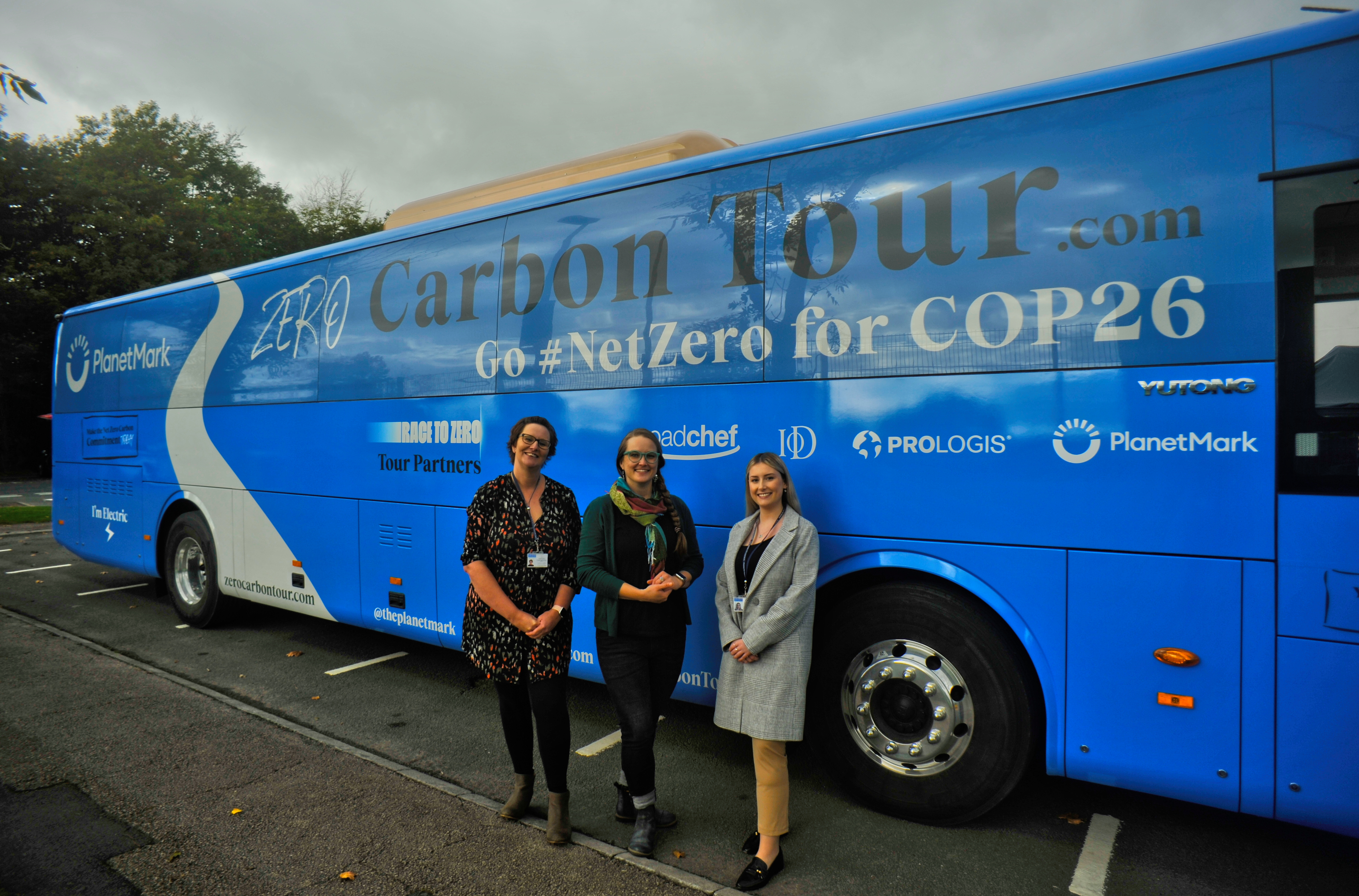Three women stood in front of a zero carbon tour coach for COP26