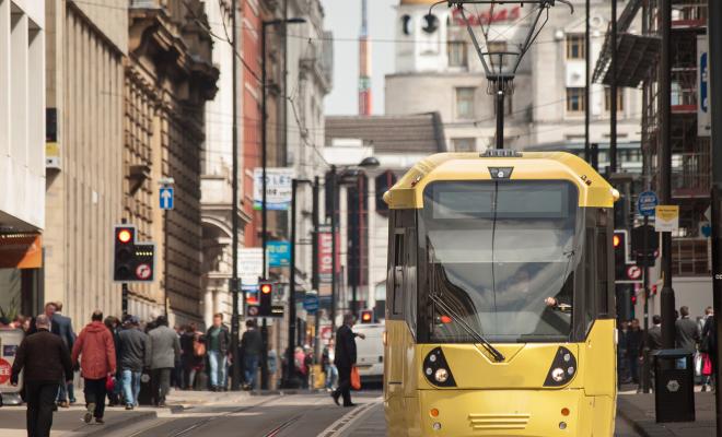 Yellow tram travels through Manchester with people on pavement.